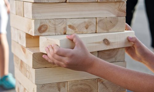 The hands of a child taking one block from a tower during a street game of giant Jenga. Outside the home. Daylighting